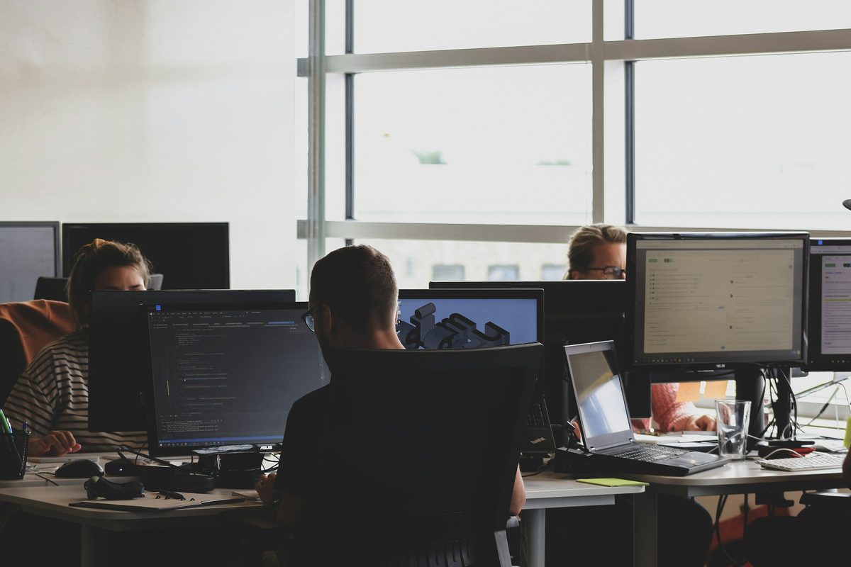 An open-plan engineering floor with developers at multi-monitor desks in front of tall windows