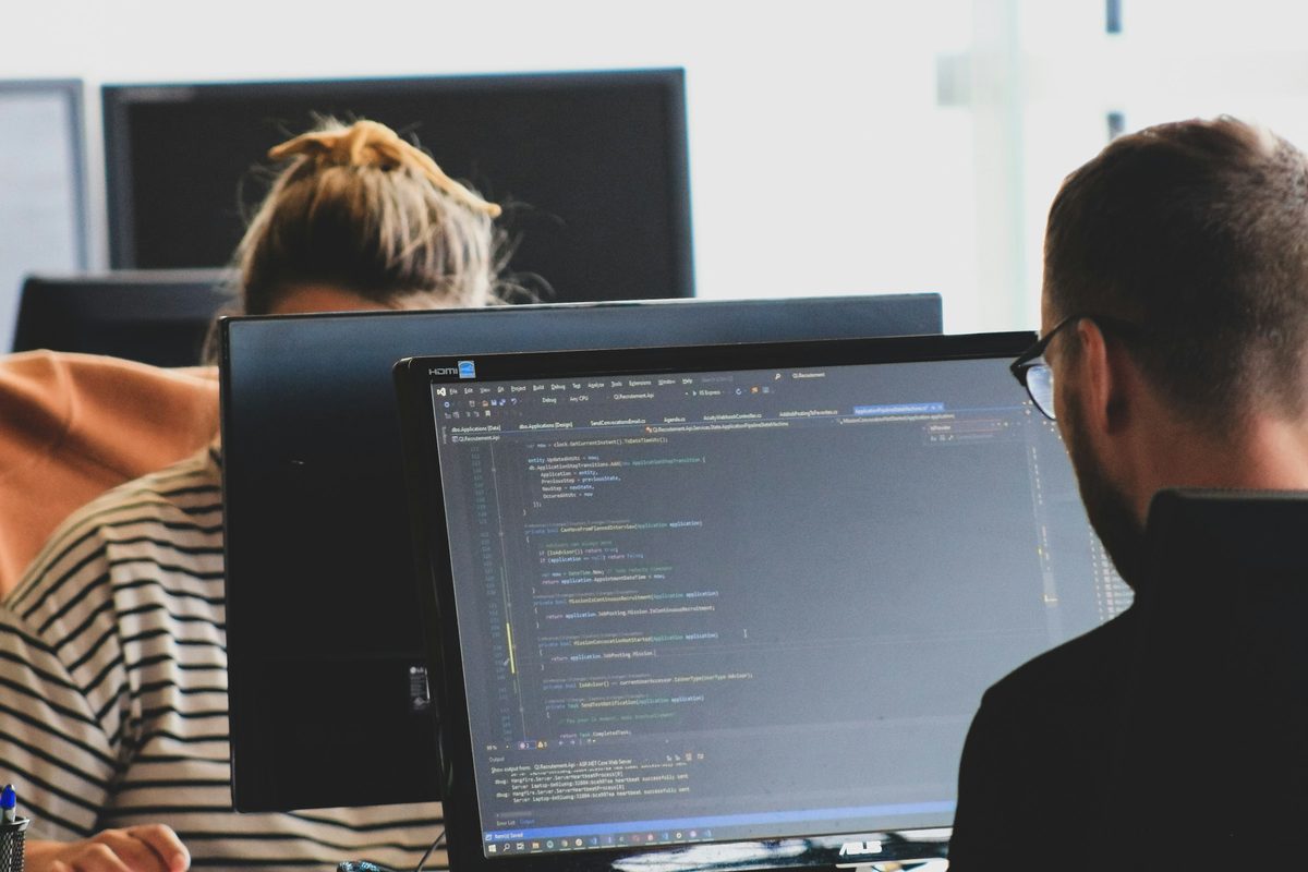 Two developers at their monitors, one working through C# code in an integrated development environment