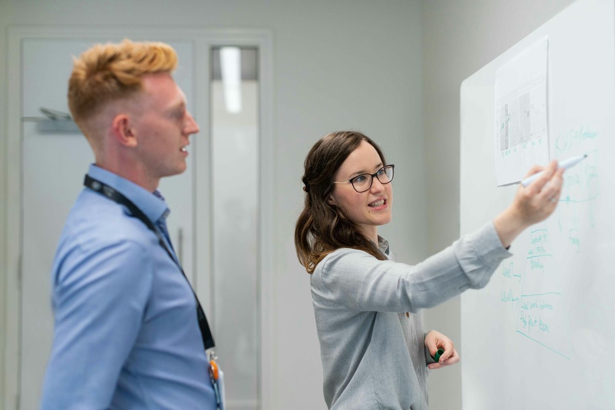 Two colleagues at a whiteboard, one pointing out levels on a hand-drawn diagram while the other listens